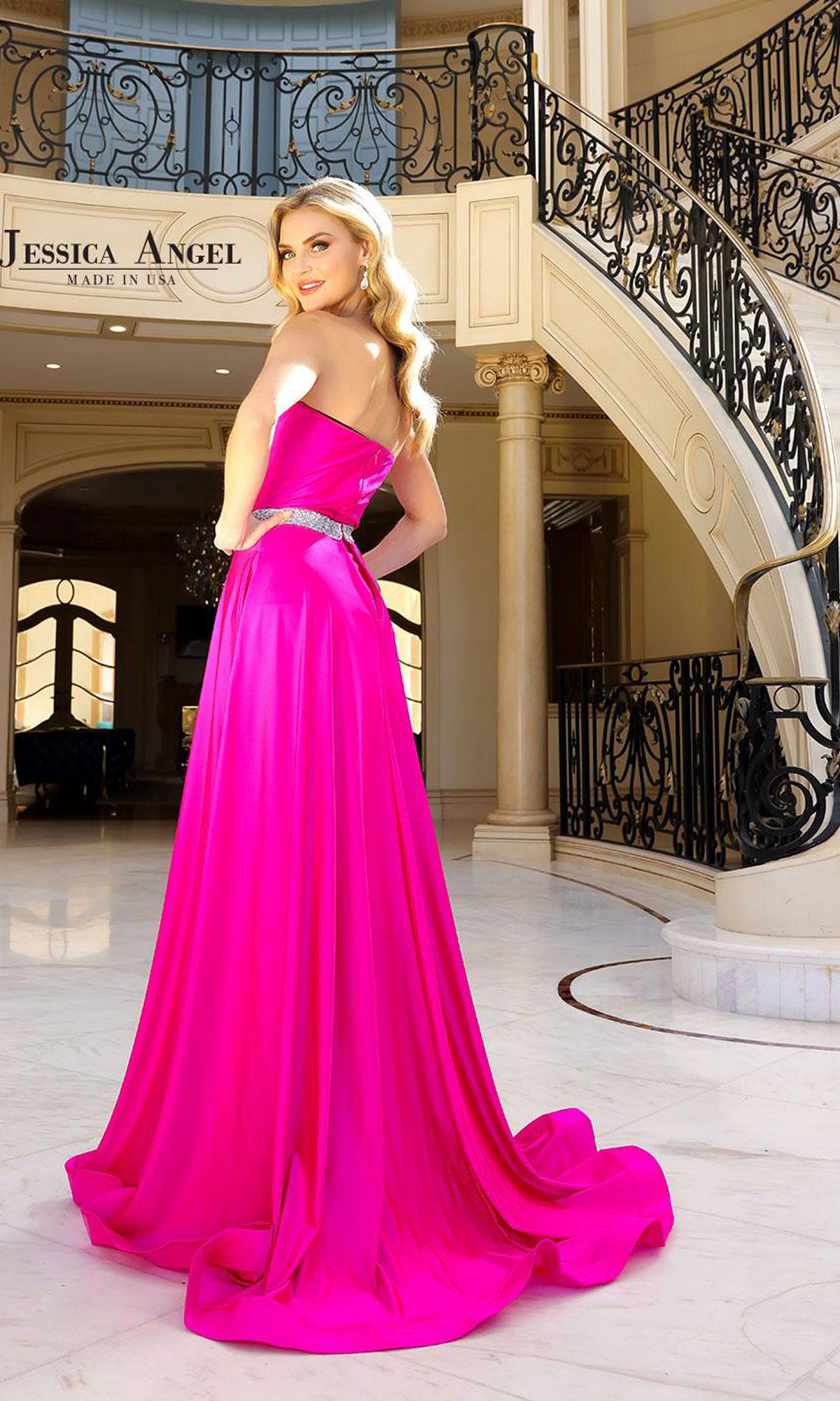 A woman wearing the Strapless High-Low Prom Dress: Jessica Angel 2402 in vibrant pink stands in an elegant foyer with a grand staircase and ornate railing in the background.